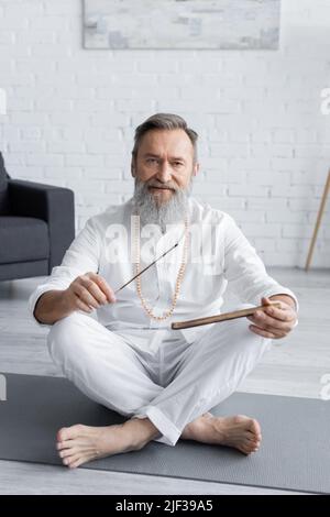 professeur de yoga barbu tenant des bâtons d'arôme tout en étant assis dans une posture facile et en regardant l'appareil photo Banque D'Images