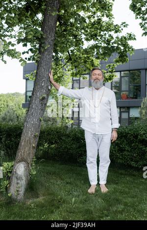 pleine longueur de gourou entraîneur touchant le tronc d'arbre tout en méditant avec les yeux fermés dehors Banque D'Images