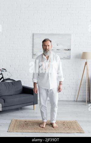 entraîneur de yoga barbu dans des vêtements blancs debout sur le tapis à la maison et regardant l'appareil photo Banque D'Images