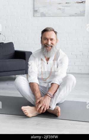 joyeux entraîneur de yoga dans les bracelets perlés assis dans la pose facile et regardant l'appareil photo Banque D'Images