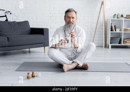 gourou de guérison barbu assis dans une posture facile avec des bâtons d'arôme près des bouteilles avec des huiles aromatisées Banque D'Images