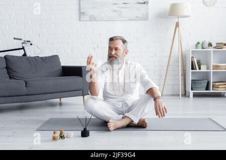 gourou de yoga senior assis avec la pierre de sélénite près des huiles parfumées et des bâtons d'arôme Banque D'Images