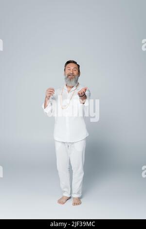 longueur complète d'entraîneur de yoga heureux dans des vêtements blancs regardant l'appareil photo et pointant avec la main sur gris Banque D'Images