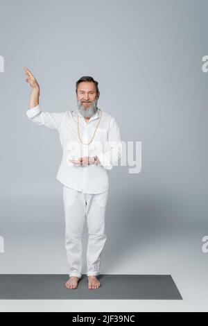 guru barbu et pieds nus en vêtements blancs qui gestent et regardent la caméra sur le gris Banque D'Images