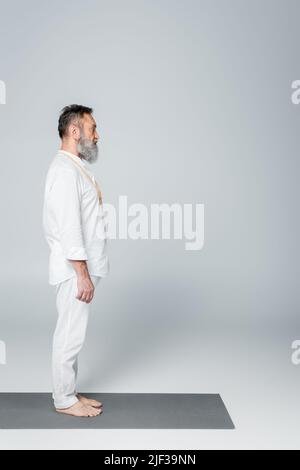 vue latérale de l'entraîneur de yoga barbu méditant dans la pose de montagne sur le tapis de yoga sur gris Banque D'Images