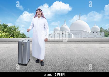 Homme arabe portant le keffiyeh debout avec une valise avec mosquée et fond bleu ciel Banque D'Images