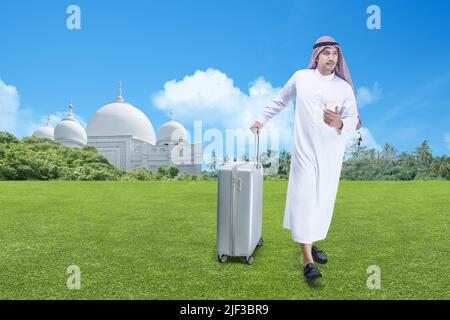 Homme arabe portant le keffiyeh tenant un téléphone portable avec une valise avec mosquée et fond bleu ciel Banque D'Images