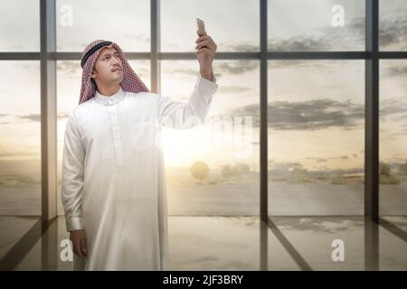 Homme arabe portant le keffiyeh regardant le téléphone mobile à l'intérieur de la pièce avec verre de fenêtre et coucher de soleil ciel fond Banque D'Images