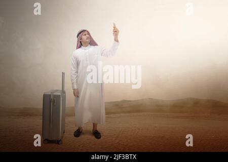 Homme arabe portant du keffiyeh regardant un téléphone portable avec une valise pour voyager dans le désert Banque D'Images
