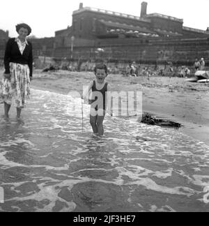 1953, historique, une mère avec son jeune fils dans les eaux peu profondes du bord de mer, avec le petit garçon debout tenant la corde attachée à son bateau en bois jouet qu'il tire à travers l'eau, Margate, Kent, Angleterre, Royaume-Uni. Banque D'Images