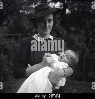 1950s, historique, debout à l'extérieur dans un jardin, une femme élégamment habillée, portant un chapeau, tenant son bébé nouvellement baptisé, Angleterre, Royaume-Uni. Banque D'Images