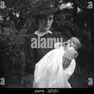 1950s, historique, debout à l'extérieur dans un jardin, une femme élégamment habillée, portant un chapeau, tenant son bébé nouvellement baptisé, Angleterre, Royaume-Uni. Banque D'Images