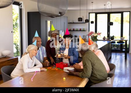 Des aînés multiraciaux portant des chapeaux de fête regardant une femme tenant des cupcakes d'anniversaire à la table Banque D'Images