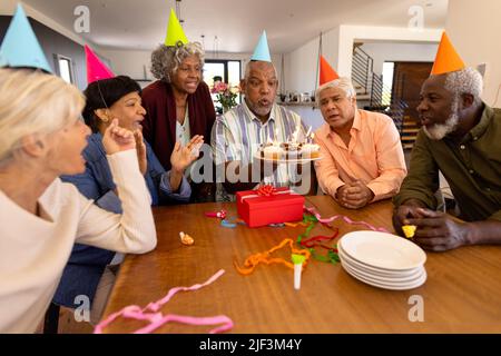 Amis séniors multiraciaux portant des chapeaux de fête regardant l'homme soufflant des bougies dans la maison de soins infirmiers Banque D'Images