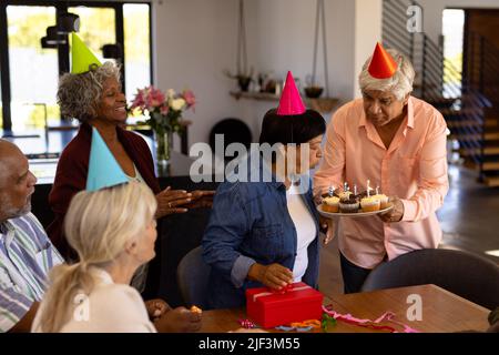 Femme sénior multiraciale soufflant des bougies d'anniversaire sur des cupcakes tenus par un ami dans une maison de soins Banque D'Images