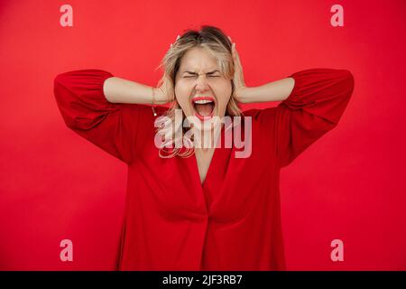 Portrait d'une femme criant haut dans une tenue rouge avec un maquillage brillant parfait tenant sur la tête par les mains isolées sur fond rouge. Perte de contrôle Banque D'Images