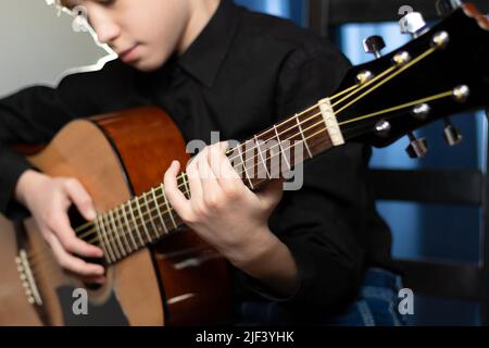 Un adolescent dans une chemise noire joue de la guitare acoustique lors d'un concert sur fond bleu. Passe-temps. Instruments de musique. Mise au point sélective. Portrait Banque D'Images