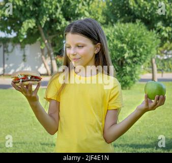 Une adolescente choisit entre un hamburger et une pomme en plein air dans le parc. Banque D'Images