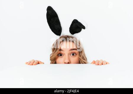 Drôle jeune femme blonde portant des oreilles de lapin de pâques peeking dehors de la table, fond blanc Banque D'Images