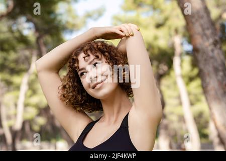 Joyeuse femme à tête rouge portant un soutien-gorge de sport noir debout sur le parc de la ville, en plein air étirant ses bras. Femme qui fait de l'entraînement d'étirement d'échauffement tout en looki Banque D'Images