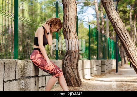 Jeune femme à tête rouge portant des vêtements de sport sur le parc de la ville, à l'extérieur après avoir fait du jogging, attrapant sa respiration, touchant son cou. Banque D'Images