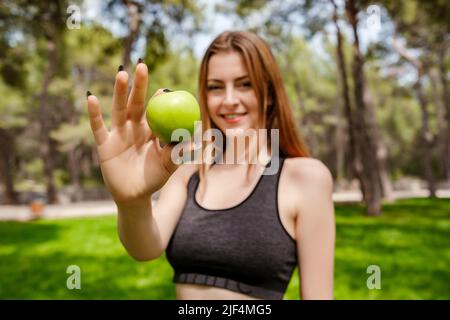 Heureuse femme sportive portant un soutien-gorge de sport debout sur le parc de la ville, en plein air souriant, tenant une pomme et regardant la caméra. Sports et santé en plein air Banque D'Images