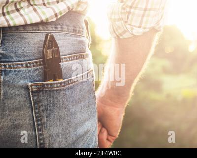 Beau homme en vêtements de travail, tenant des outils dans ses mains contre le fond des arbres, le ciel bleu et le coucher du soleil. Vue de l'arrière, gros plan, extérieur. Banque D'Images