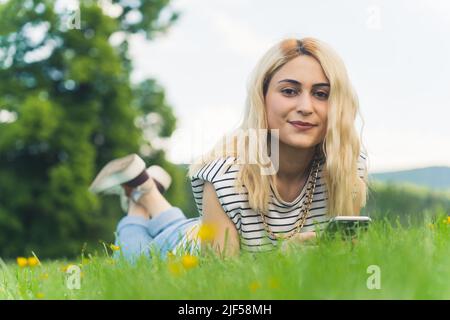 Jolie jeune fille caucasienne sur l'herbe tenant un téléphone et regardant l'appareil photo. Plein plan extérieur. Photo de haute qualité Banque D'Images