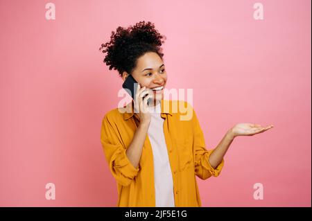 Conversation téléphonique par téléphone portable. Heureuse joyeuse excitée africaine jeune femme américaine, a une conversation téléphonique agréable, expression du visage heureux, se tient sur un fond rose isolé, la main gestante, souriant Banque D'Images