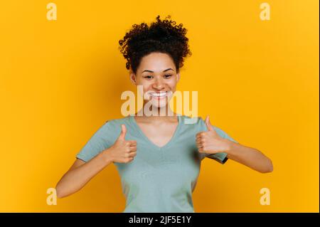 Positive adorable jeune afro-américaine femme aux cheveux bouclés, vêtue d'un t-shirt de base, montrant les pouces vers le haut geste, regarde l'appareil photo, debout sur un arrière-plan orange isolé, souriant heureusement Banque D'Images