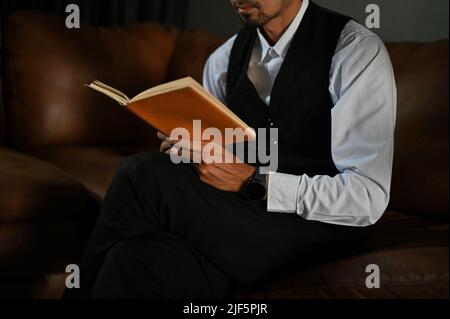 Image rognée, Un homme d'affaires asiatique professionnel et élégant en costume habillé, assis sur un canapé en cuir et lisant un livre. Banque D'Images