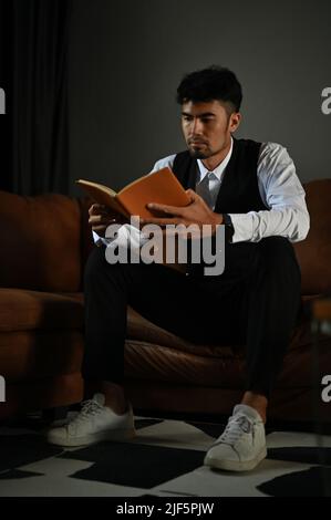 Élégant et élégant homme d'affaires asiatique ou directeur exécutif masculin se trouve sur un canapé en cuir et lit un livre. Banque D'Images