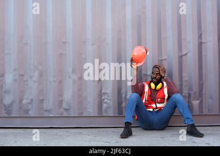 Technicien ou ingénieur afro-américain. S'asseoir près d'un récipient et avoir l'air fatigué et endormi ou sans emploi. Logistique concept de transport de fret industriel. Banque D'Images