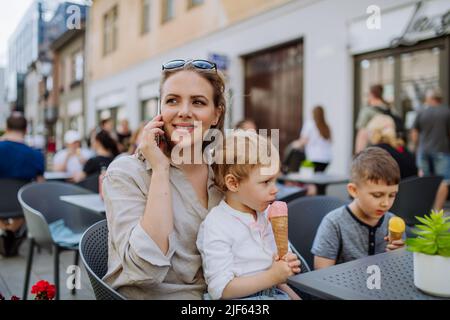 Une jeune mère avec ses enfants mangeant de la glace dans un café à l'extérieur dans la rue en été. Banque D'Images