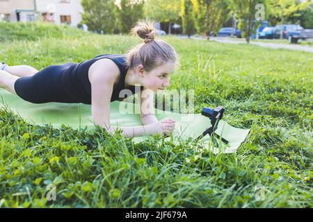 Jeune femme blonde de forme pratiquant la planche sur le tapis de yoga et l'enregistrement vidéo tutoriel sur la caméra extérieure le jour ensoleillé d'été. FIT gen z girl faisant du yoga sur l'herbe dans le parc. Mode de vie sain, cours de yoga en ligne, concept de sport et d'activité physique Banque D'Images