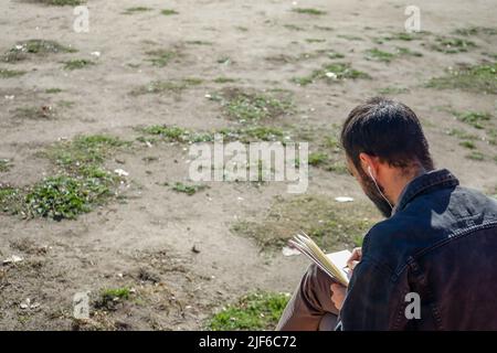 Homme écrivant ou dessinant sur un carnet dans un parc. Banque D'Images