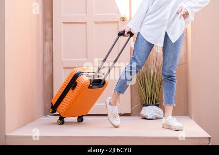 Jeune femme en solo se tenant debout avec la porte géorgienne alors qu'elle remorque sa valise orange vif remplie de techniques de travail à distance, en mettant des billets d'avion dans sa poche gauche Banque D'Images