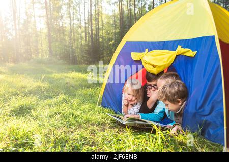 Quatre enfants d'âge différent lisant un livre couché ensemble dans le camping tente dans une forêt d'été pendant les vacances d'été Banque D'Images