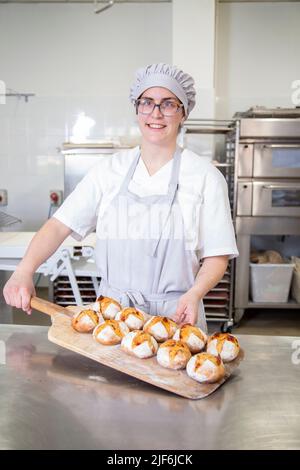 Boulanger féminine gaie en uniforme avec abondance de petit pain cuit regardant l'appareil photo à la table dans la boulangerie avec des collègues Banque D'Images