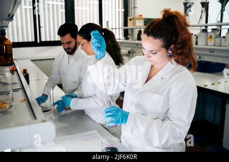 Chercheuse concentrée en uniforme médical, debout près de collègues tout en versant du liquide chimique coloré de la pipette dans le travail en flacon de verre Banque D'Images