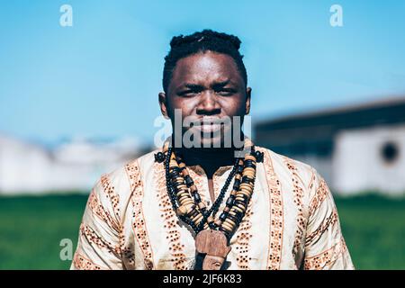 Homme noir en chemise africaine ornementale et colliers authentiques frognant et regardant la caméra sur un fond flou de ciel bleu et de bâtiments de ferme dans Banque D'Images
