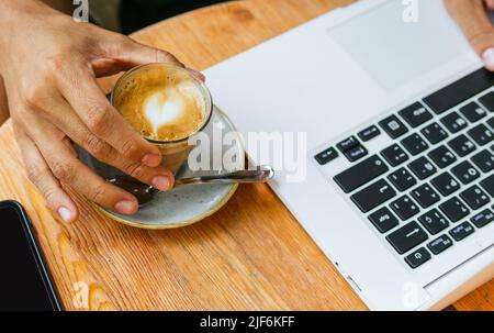 Un indépendant anonyme de sexe masculin boit du café tout en travaillant à distance sur un netbook moderne dans un café Banque D'Images