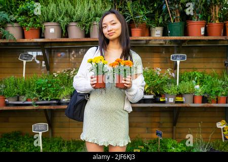 Femelle asiatique positive avec des marigolds fleuris dans des pots regardant l'appareil photo tout en se tenant près des étagères avec des fleurs variées sur le marché Banque D'Images