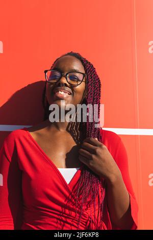 Jeune femme afro-américaine gaie dans une tenue tendance et des lunettes regardant l'appareil photo tout en se tenant sur fond rouge Banque D'Images