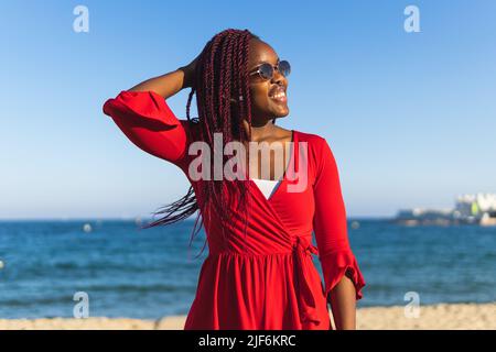 Portrait de la jeune femme afro-américaine en robe rouge tendance et tresses en regardant loin en se tenant à la plage Banque D'Images