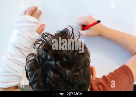 D'en haut petit garçon non reconnaissable court avec bras cassé dans le bandage en plâtre dessin image avec marqueur rouge tout en étant assis à la table le week-end Banque D'Images