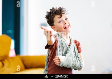 Un garçon heureux avec un bras cassé dans une élingue regardant l'appareil photo avec le sourire et faisant un geste de bienvenue sur un arrière-plan flou de la salle de séjour à la maison Banque D'Images