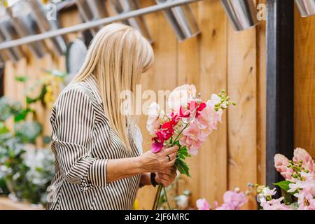 Vue latérale d'une femme âgée dans des vêtements élégants et décontractés avec de longs cheveux blonds en choisissant des fleurs fraîches pour le bouquet tout en travaillant dans le magasin Banque D'Images