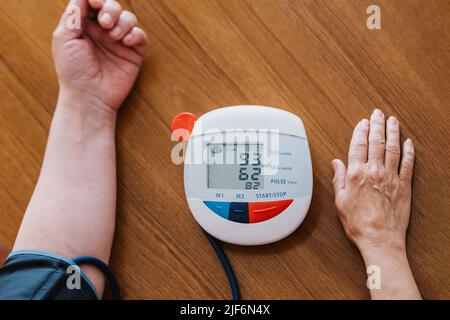 De dessus de la récolte femelle mature non reconnaissable mesurant la tension artérielle avec le sphygmomanomètre électronique moderne tout en étant assis à une table en bois Banque D'Images