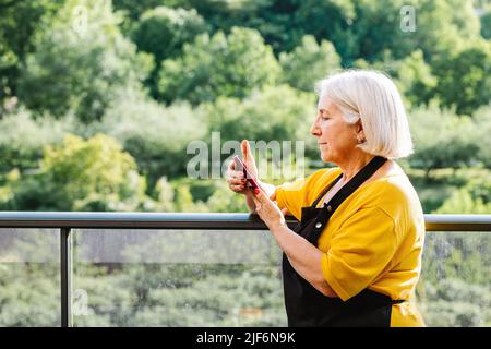 Femme sénior positive dans des vêtements décontractés se reposant sur un balcon et utilisant un téléphone portable Banque D'Images